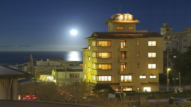 A multi-story building with illuminated windows and balconies, a visible moon in the sky, and a body of water in the background. - Aji no Yado Kikuta (Higashiizu)