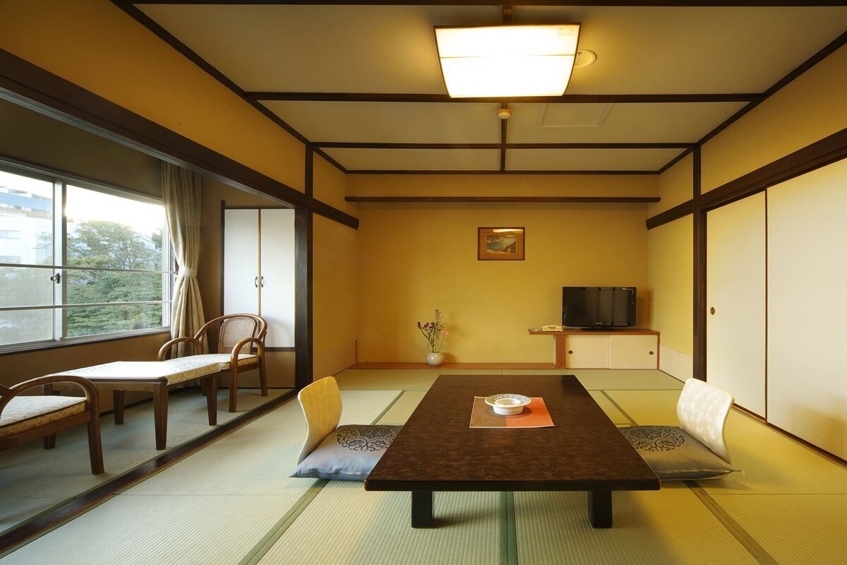 Une pièce de style japonais traditionnel, avec un sol recouvert de tatamis, une petite table basse avec un coussin, un téléviseur et des portes coulissantes.