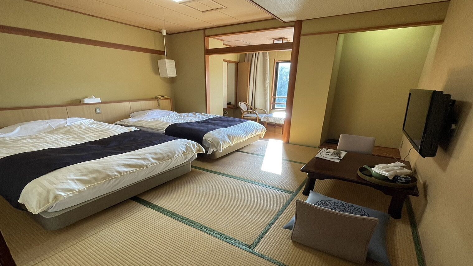 A hotel room with two beds, a small table, a chair, a TV, and a window with curtains.