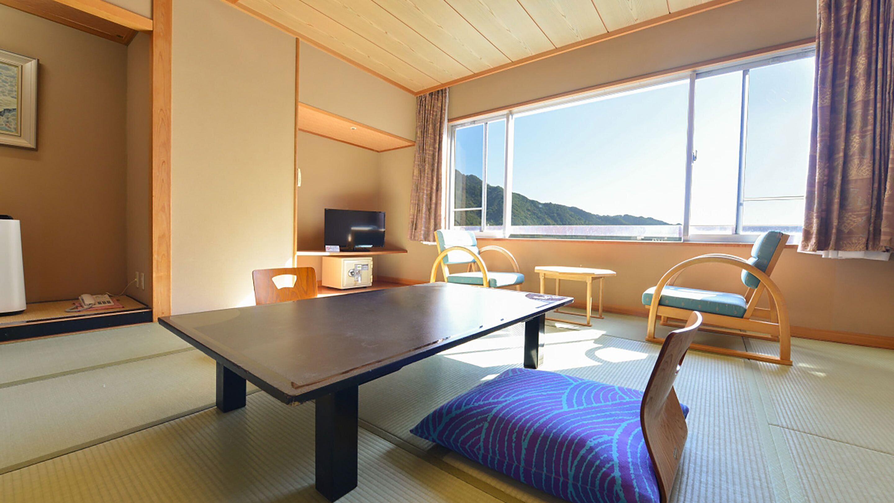 A room with tatami flooring, a low table, a television, and a view of a mountain through large windows.