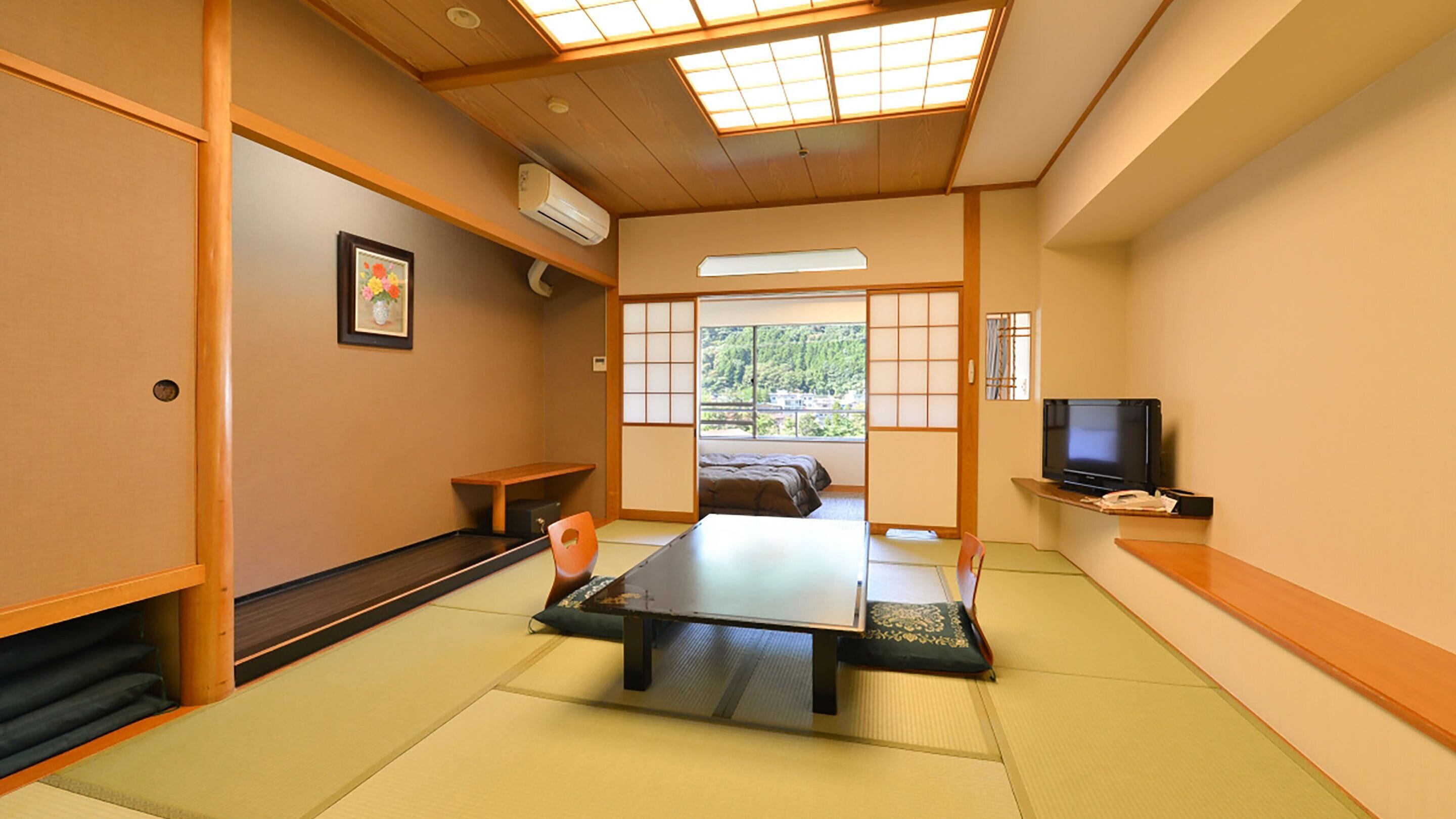 Una habitación japonesa tradicional con suelo de tatami, una mesa baja, una cama con futón, un televisor y una ventana con vistas a zonas verdes.