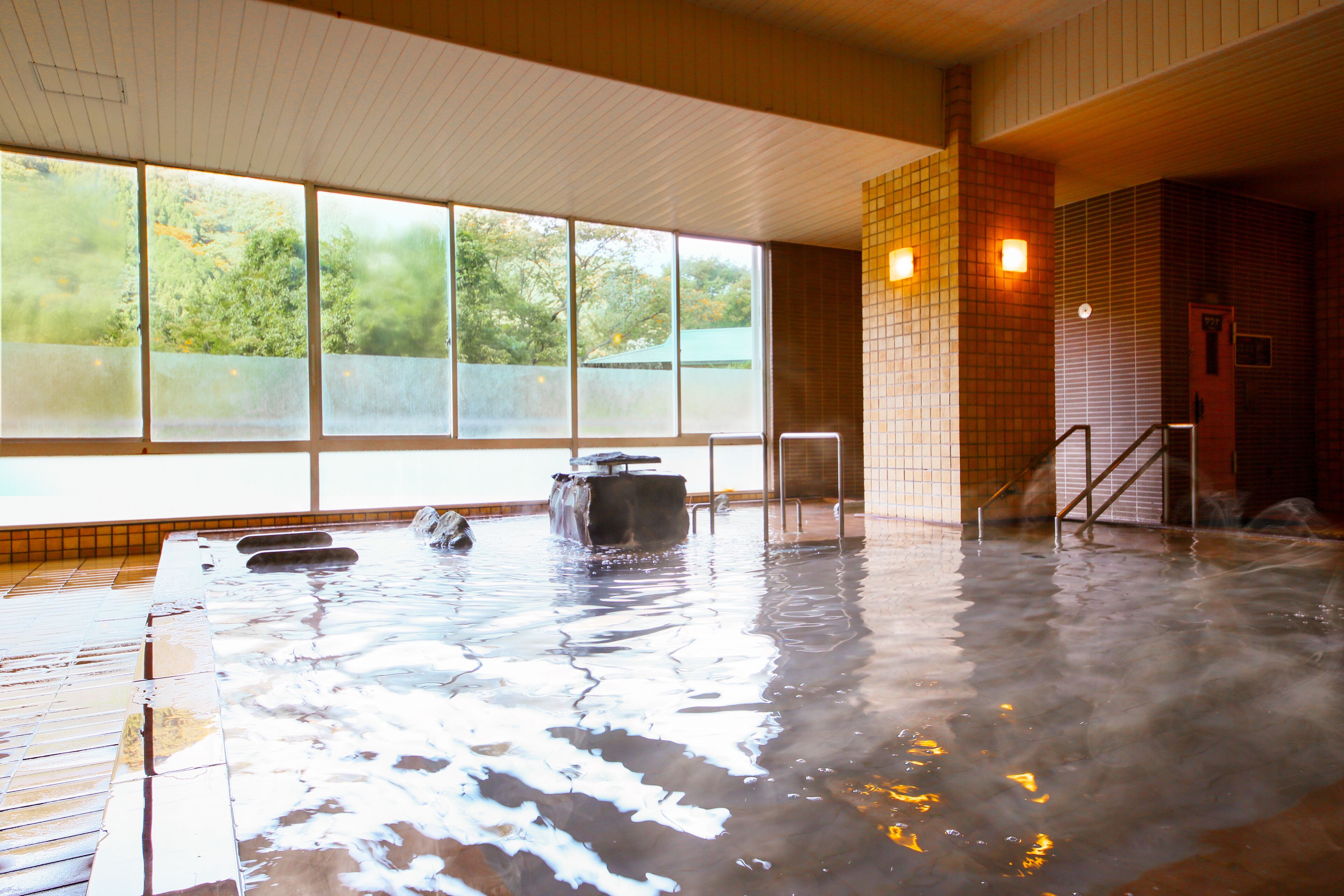 A hot spring bath with a view of trees through large windows, a tiled wall, and a staircase leading to an upper level.