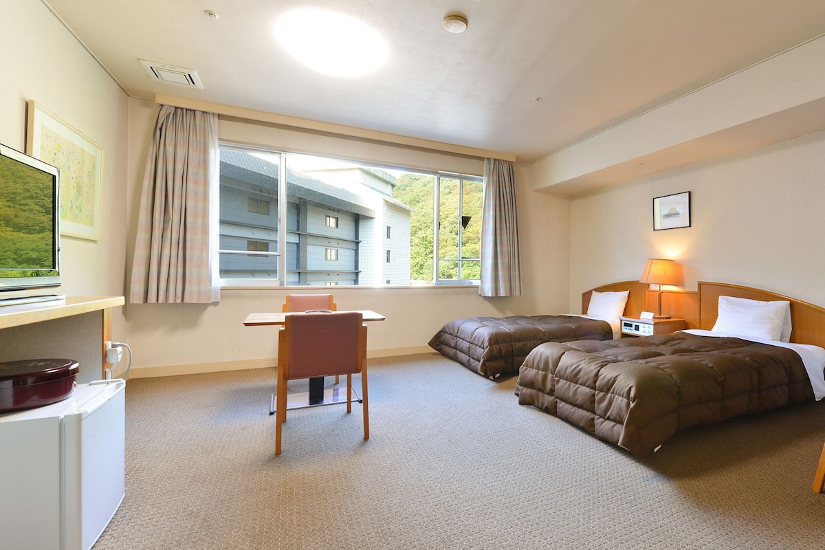 Ett hotellrum med två sängar, ett skrivbord, en stol, en TV och ett fönster med gardiner.