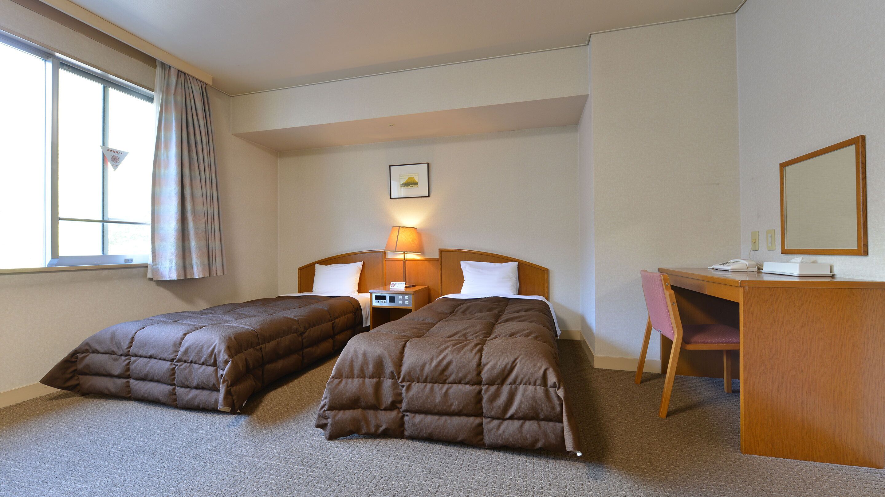 A hotel room with two single beds, a desk with a chair, a lamp, and a small picture on the wall.