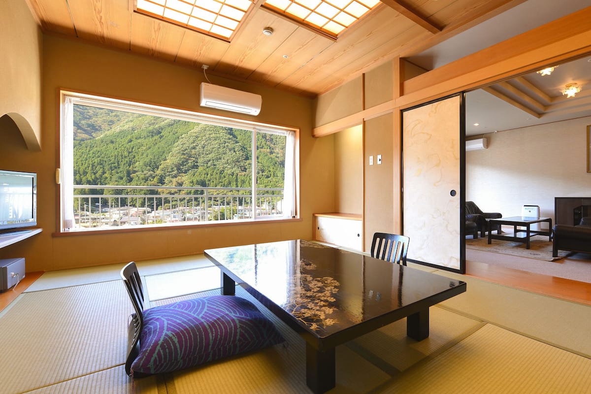 Ett traditionellt japanskt rum med tatamimattor på golvet, ett lågt bord med en kudde, en skjutdörr, en soffa och utsikt över ett skogsområde genom ett stort fönster.