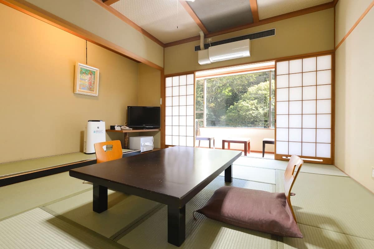 Ett traditionellt japanskt rum med tatamimattor på golvet, ett lågt bord, en tv och ett stort fönster med utsikt över träd.