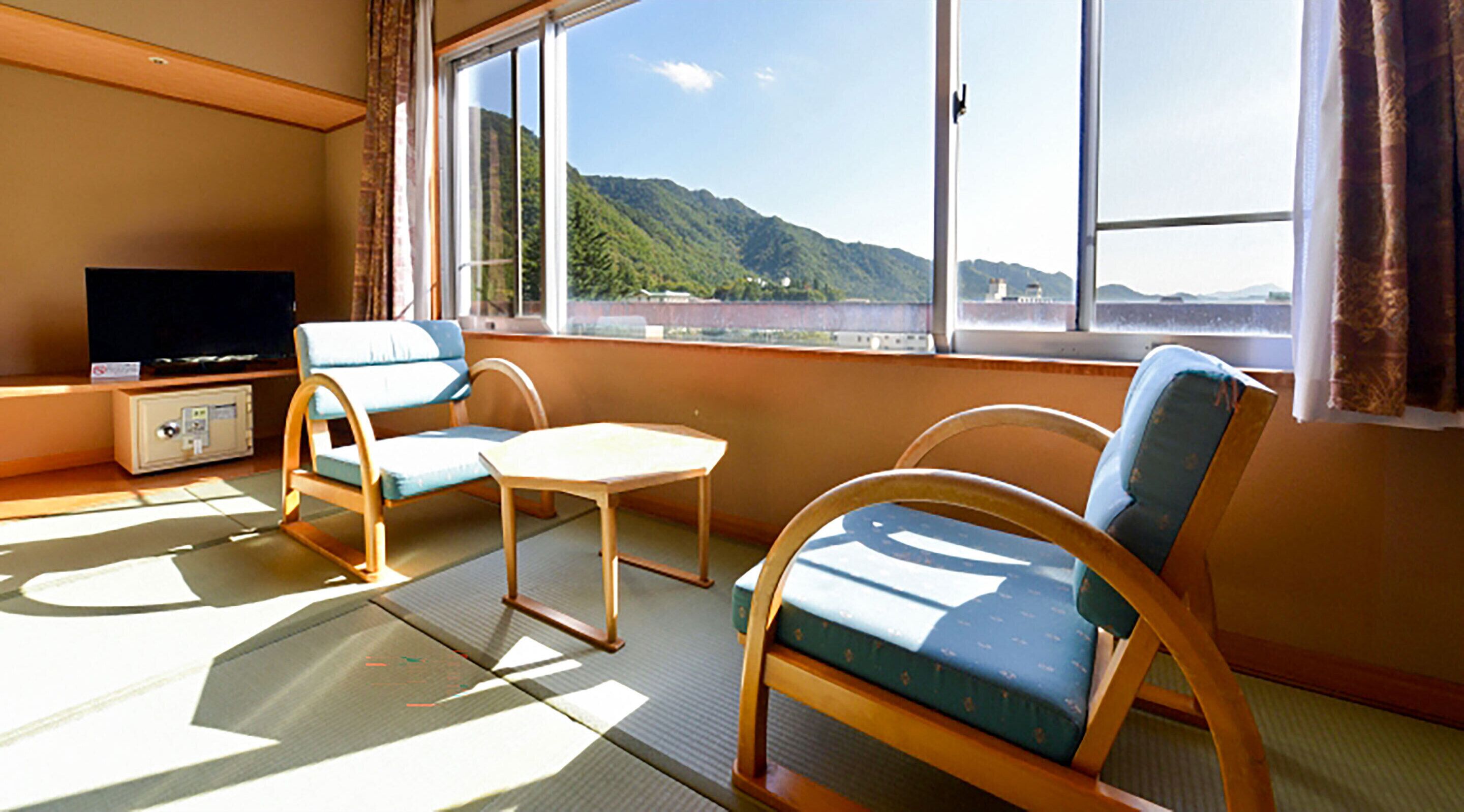 Une pièce avec un téléviseur, deux fauteuils bleus, une petite table et une vue sur une montagne, visible à travers de grandes fenêtres.