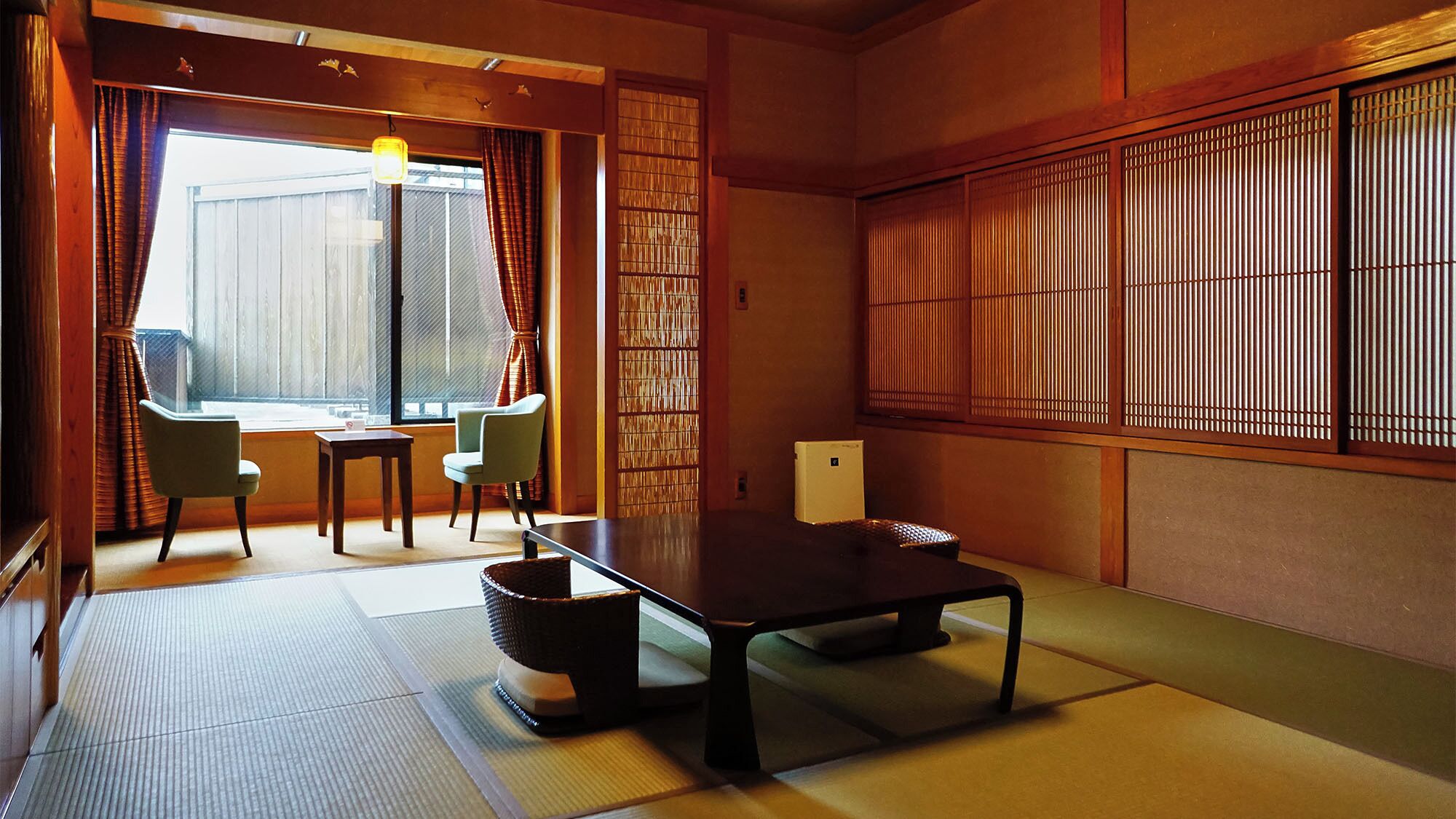 Una habitación japonesa tradicional con suelo de tatami, una mesa baja, sillas, una ventana con cortinas y puertas correderas.