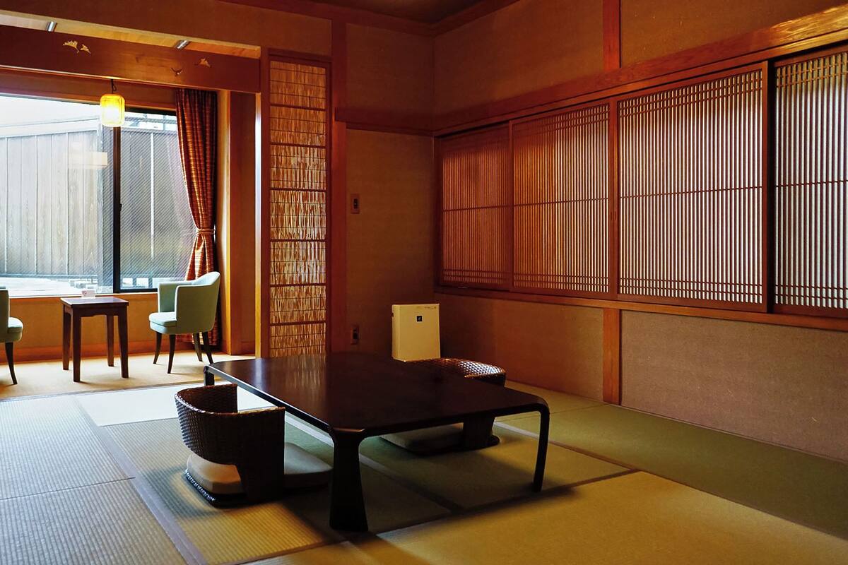 Ein traditioneller japanischer Raum mit Tatami-Böden, einem niedrigen Tisch, Stühlen, einem Fenster mit Vorhängen und Schiebetüren.