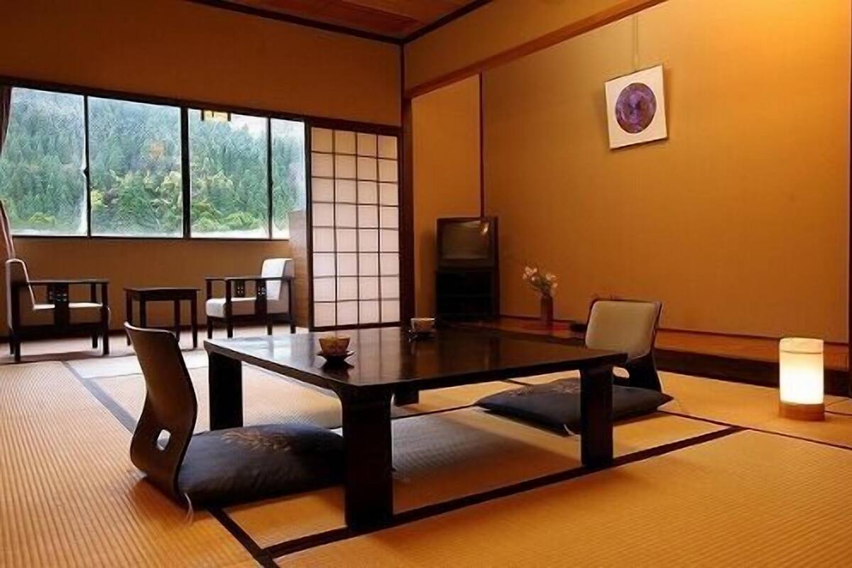Ein traditioneller japanischer Raum mit Tatami-Böden, einem niedrigen Tisch, Stühlen, einem Fernseher und einem Fenster mit Blick auf Grünfläche.