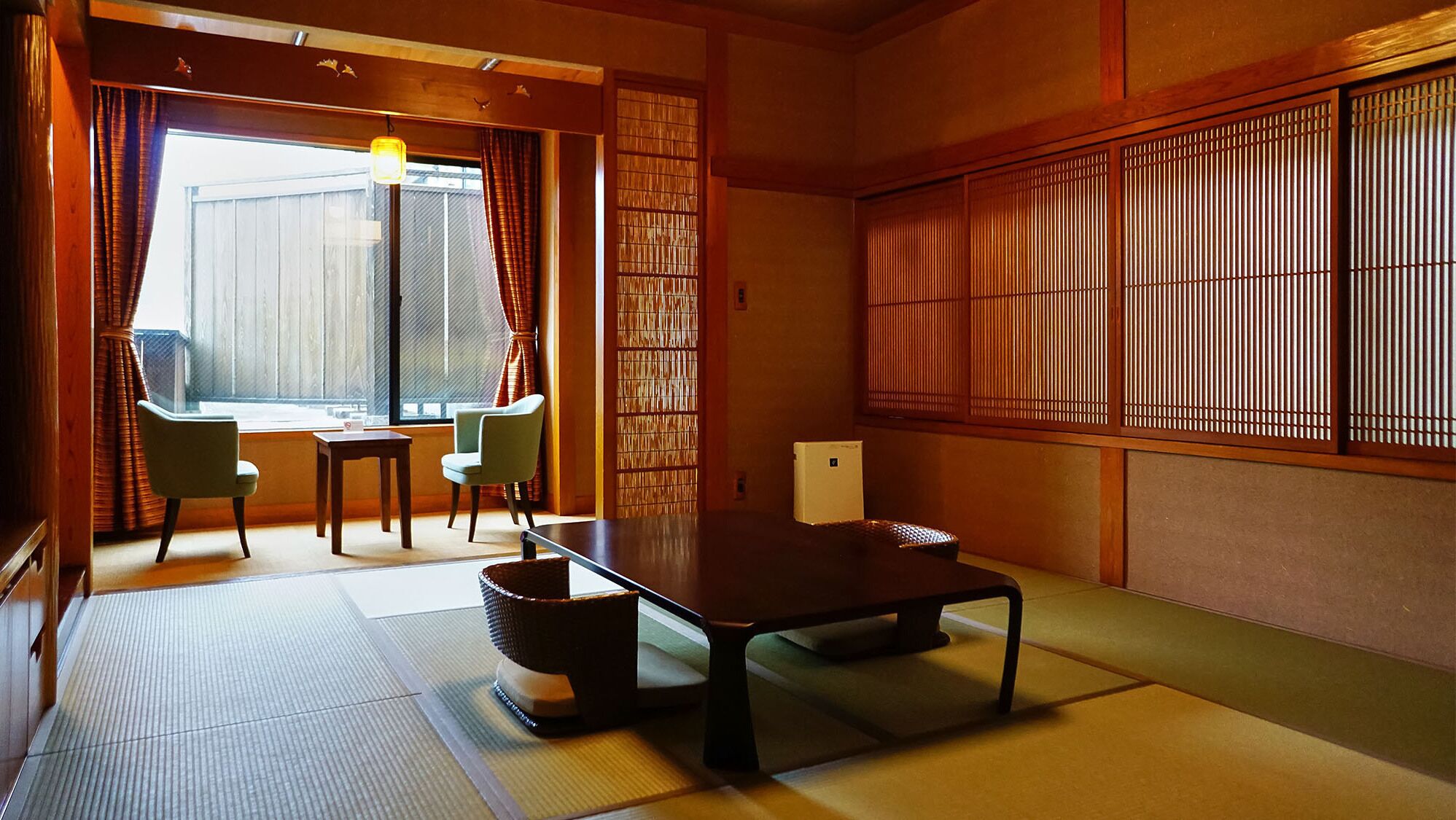Una habitación japonesa tradicional con suelo de tatami, una mesa baja, dos sillas, una mesita y una puerta corrediza con persianas verticales.