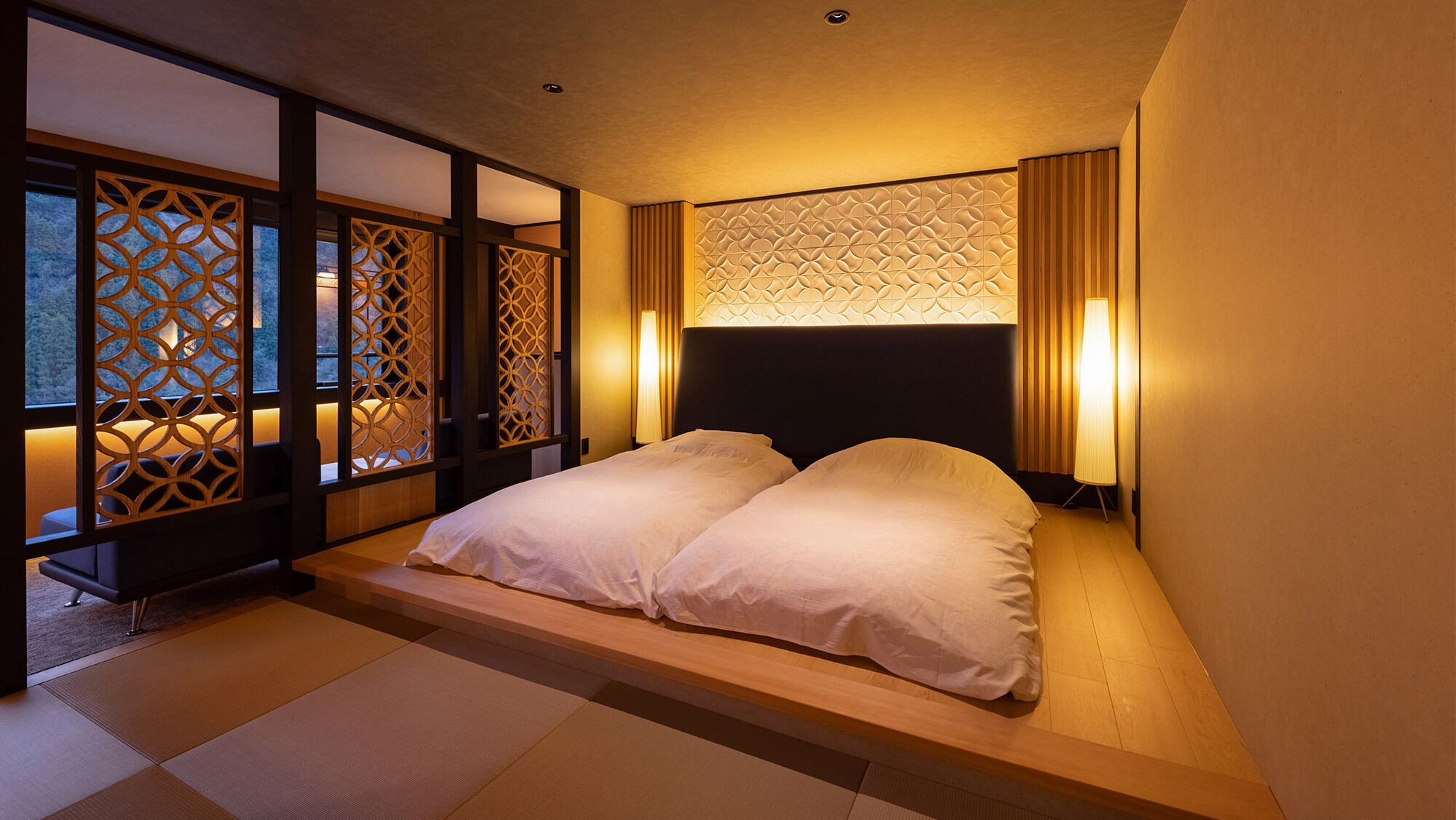 Um quarto japonês tradicional com piso de tatame, cama baixa em plataforma, portas deslizantes com padrões geométricos e cabeceira com design texturizado.