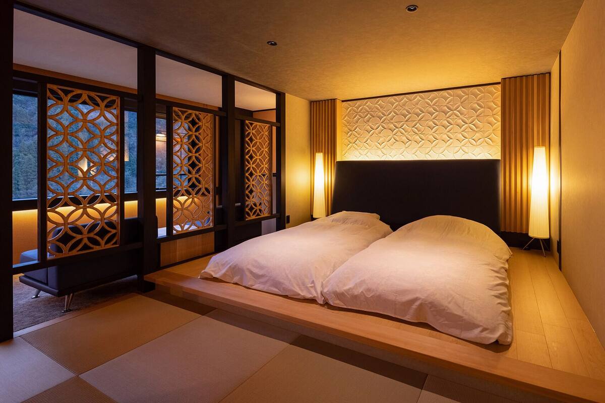 Ein traditioneller japanischer Raum mit Tatami-Boden, einer niedrigen Plattform-Liege, Schiebetüren mit geometrischen Mustern und einem Kopfteil mit strukturierter Gestaltung.