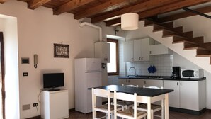 A compact kitchen with white cabinets, a refrigerator, a microwave, and a coffee maker. There is a dining table with chairs and a television on a small cabinet. - Ca' Pignoi (Garda)