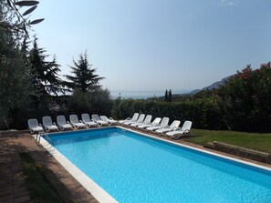 Piscina con sillas de descanso blancas dispuestas a su alrededor, rodeada de vegetación y vistas al mar.
