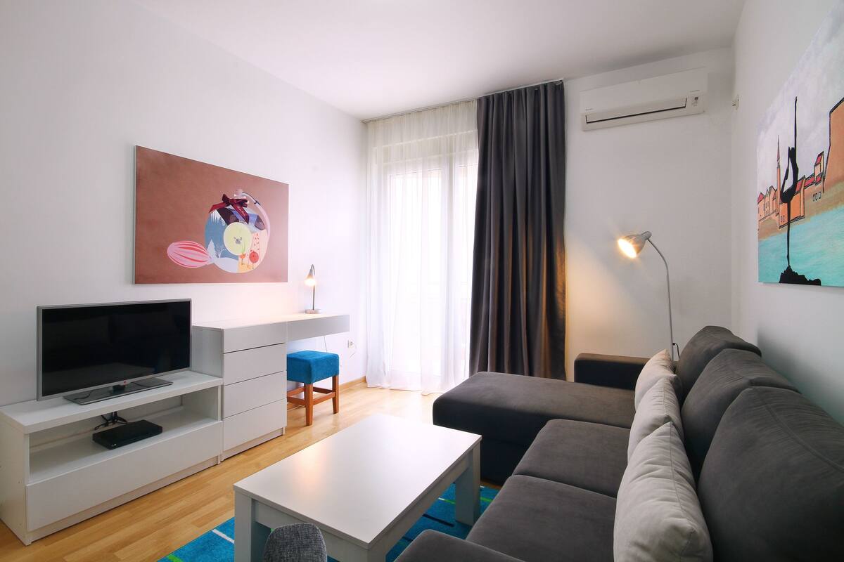 Una sala de estar moderna con un televisor de pantalla plana, muebles blancos, un reposapiés azul, un sofá gris y obras de arte en las paredes.