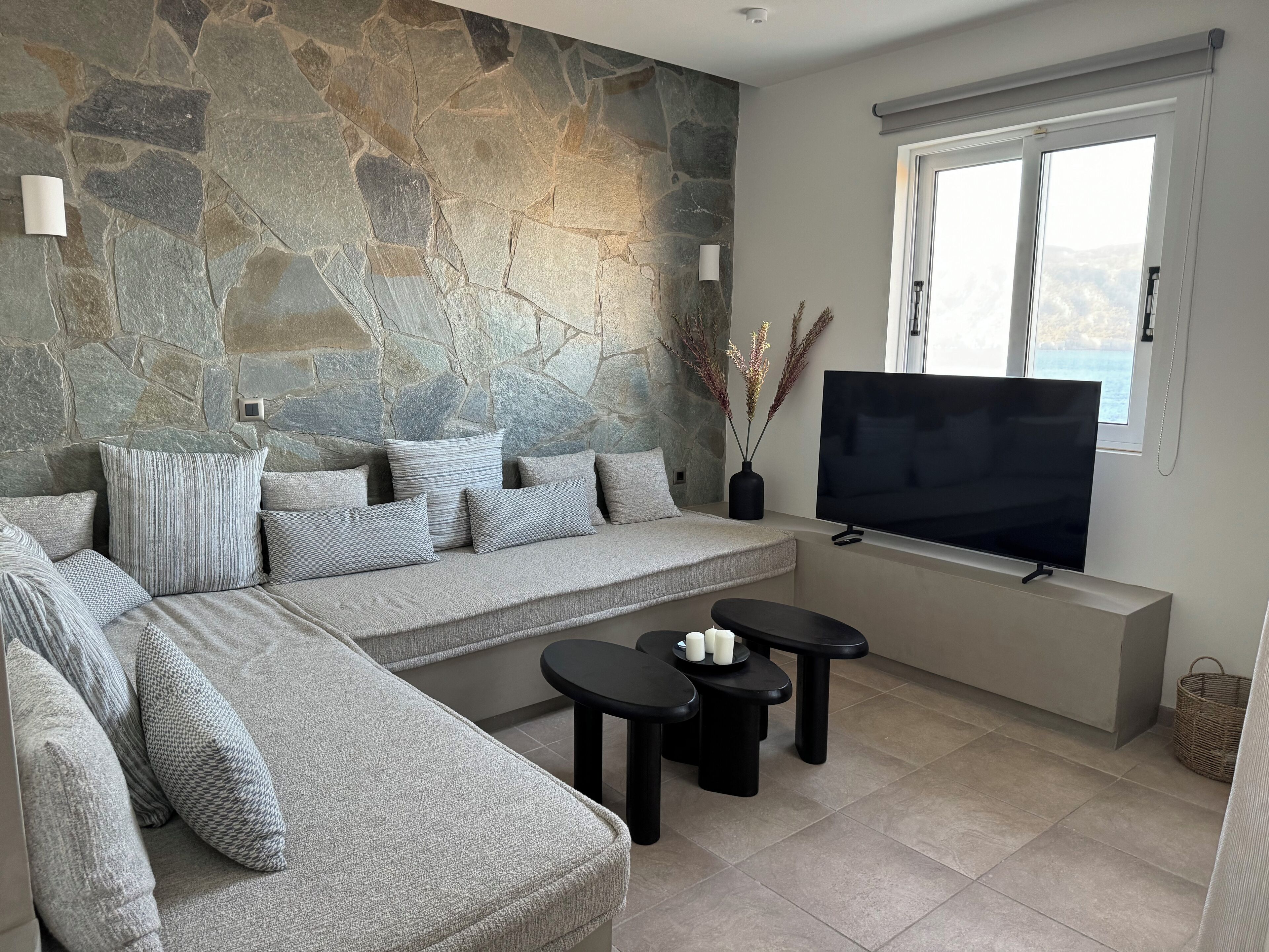 Uma sala de estar moderna com uma parede de destaque em pedra, uma grande televisão de tela plana, um sofá seccional cinza com almofadas estampadas e uma pequena mesa preta com velas.