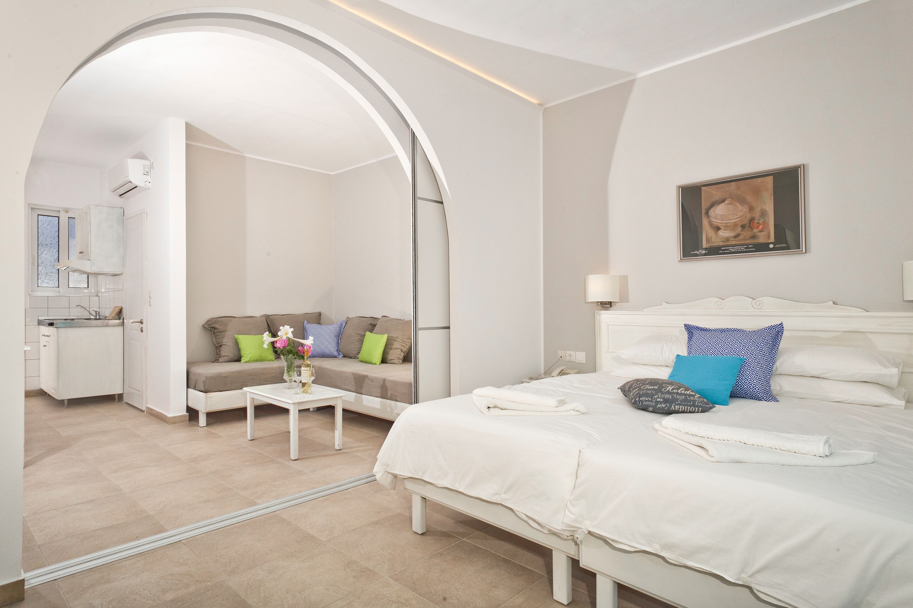 A modern hotel room with a bed, a sofa, a small table with flowers, and a bathroom visible through an archway.