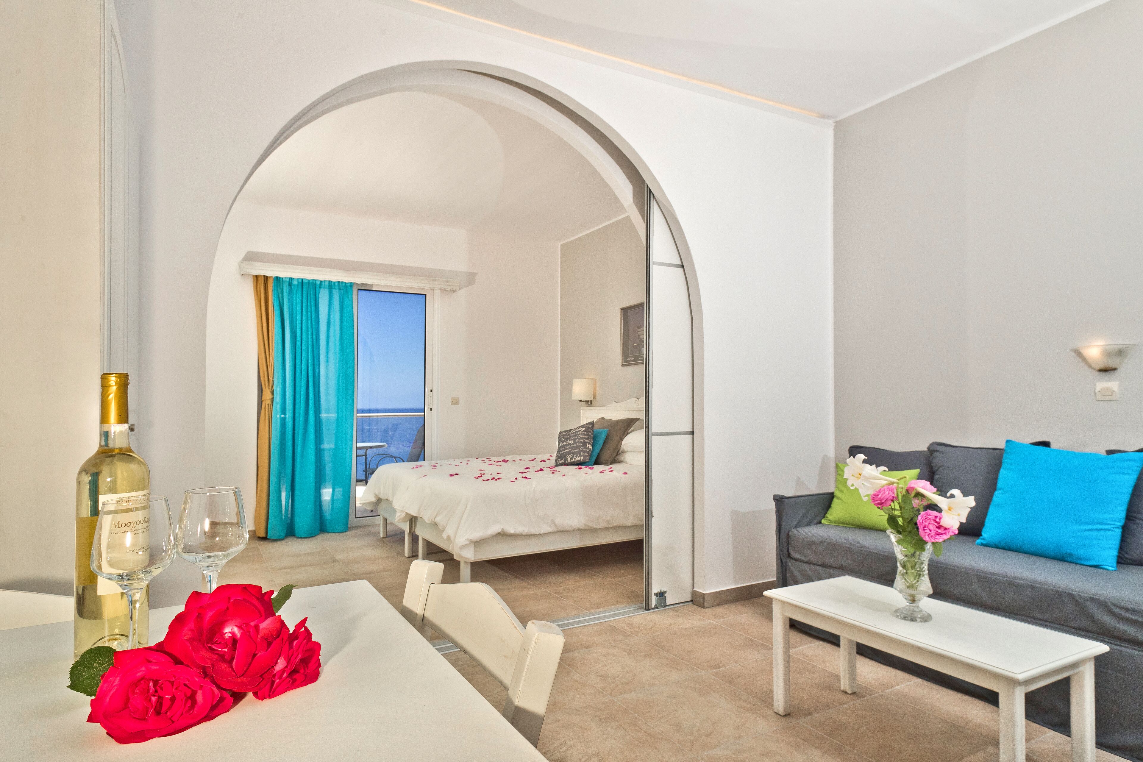 Une chambre d’hôtel avec un lit, un canapé, une table à manger décorée de roses rouges, un vase rempli de fleurs et une bouteille de vin.