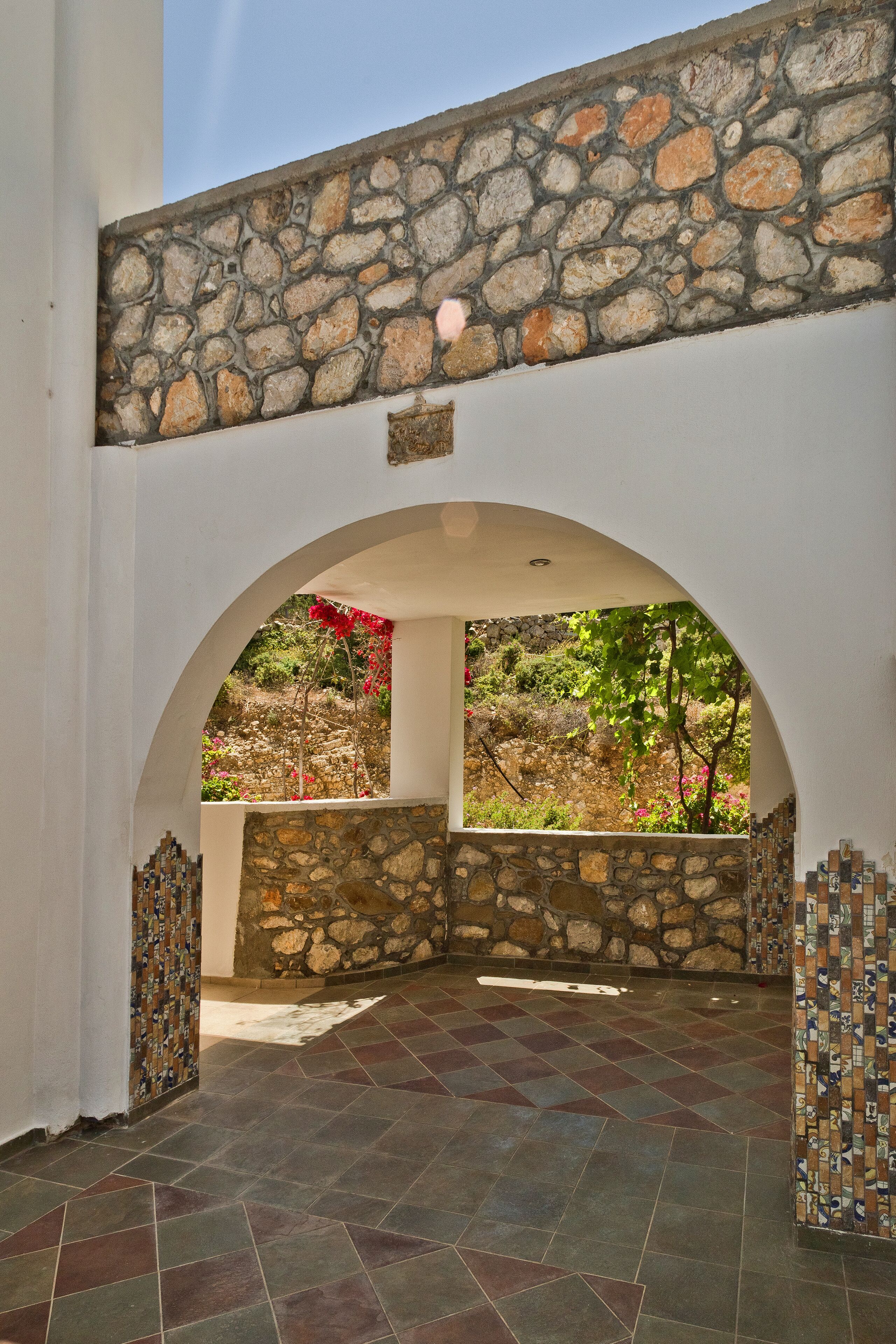 An arched stone entrance with a tiled floor, leading to a stone wall and a view of a garden.