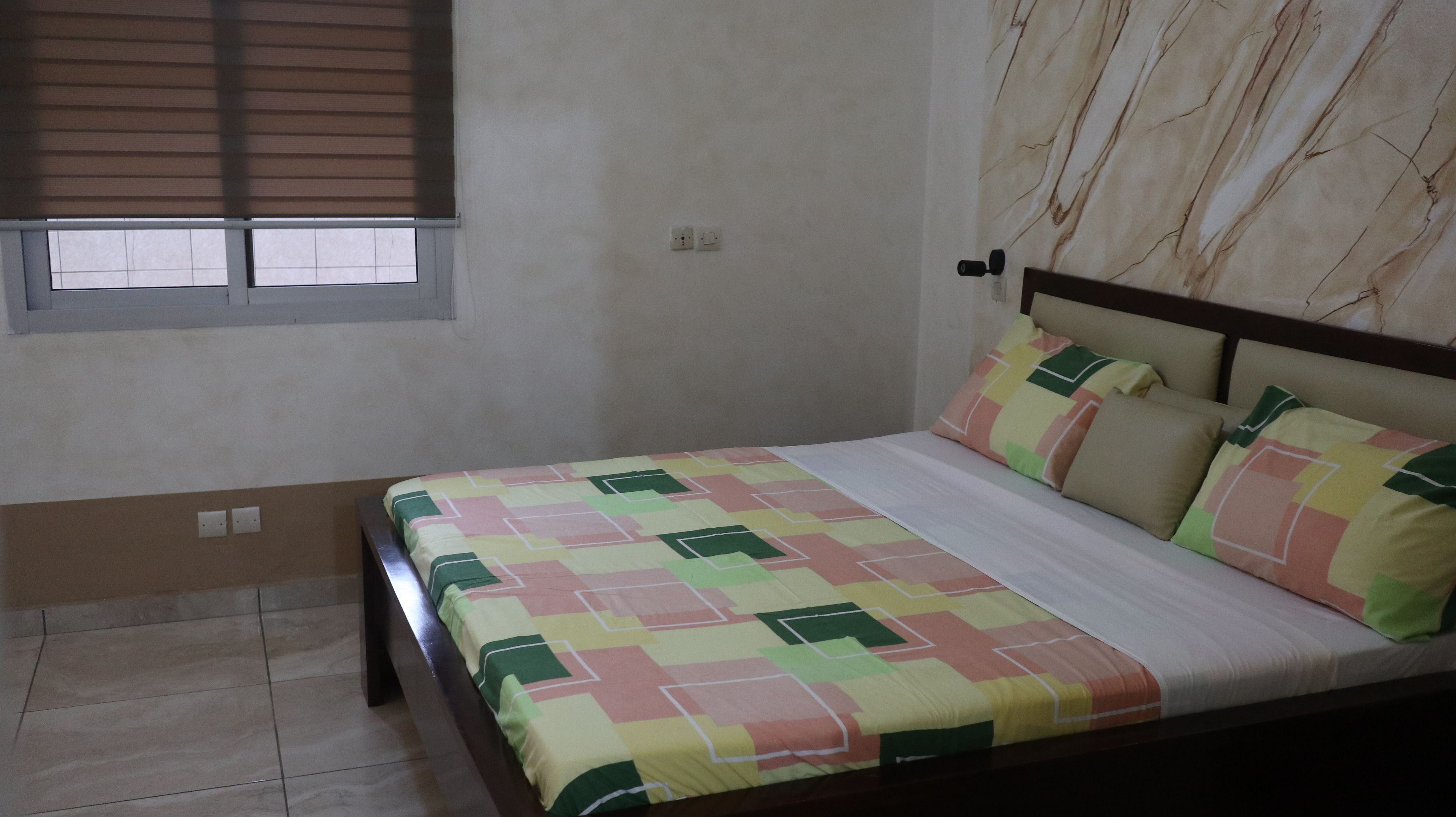 A bedroom with a bed, a window with blinds, a marble-patterned wall, and a tiled floor.
