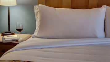 A neatly made bed with white linens, a bedside table with a lamp, a glass of red wine, and a book.