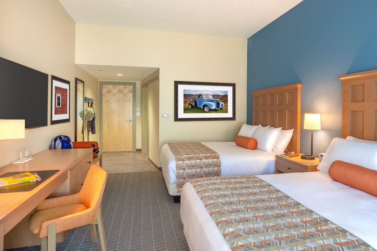 A hotel room with two beds, a desk, and a chair. There is a framed picture on the wall and a lamp on the nightstand.