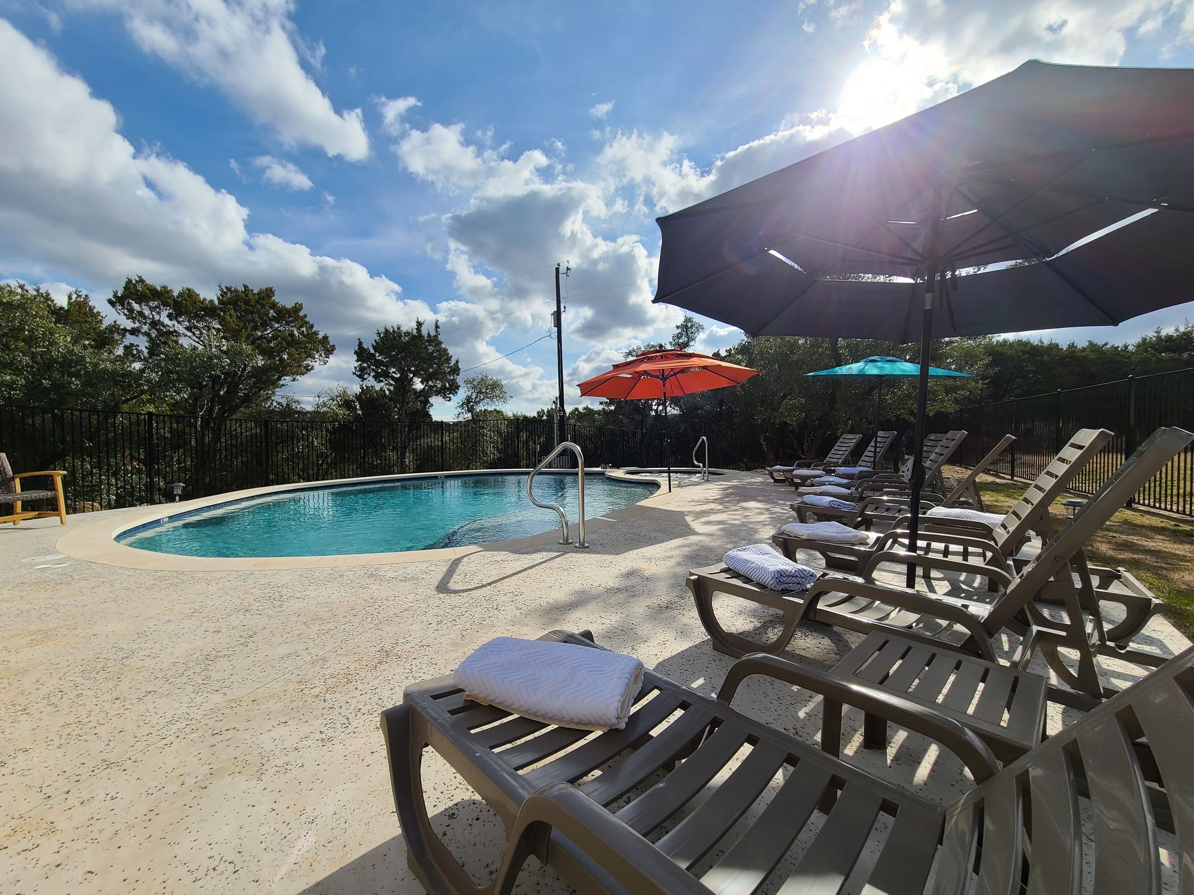 A pool area with lounge chairs and umbrellas, surrounded by a fence and trees.