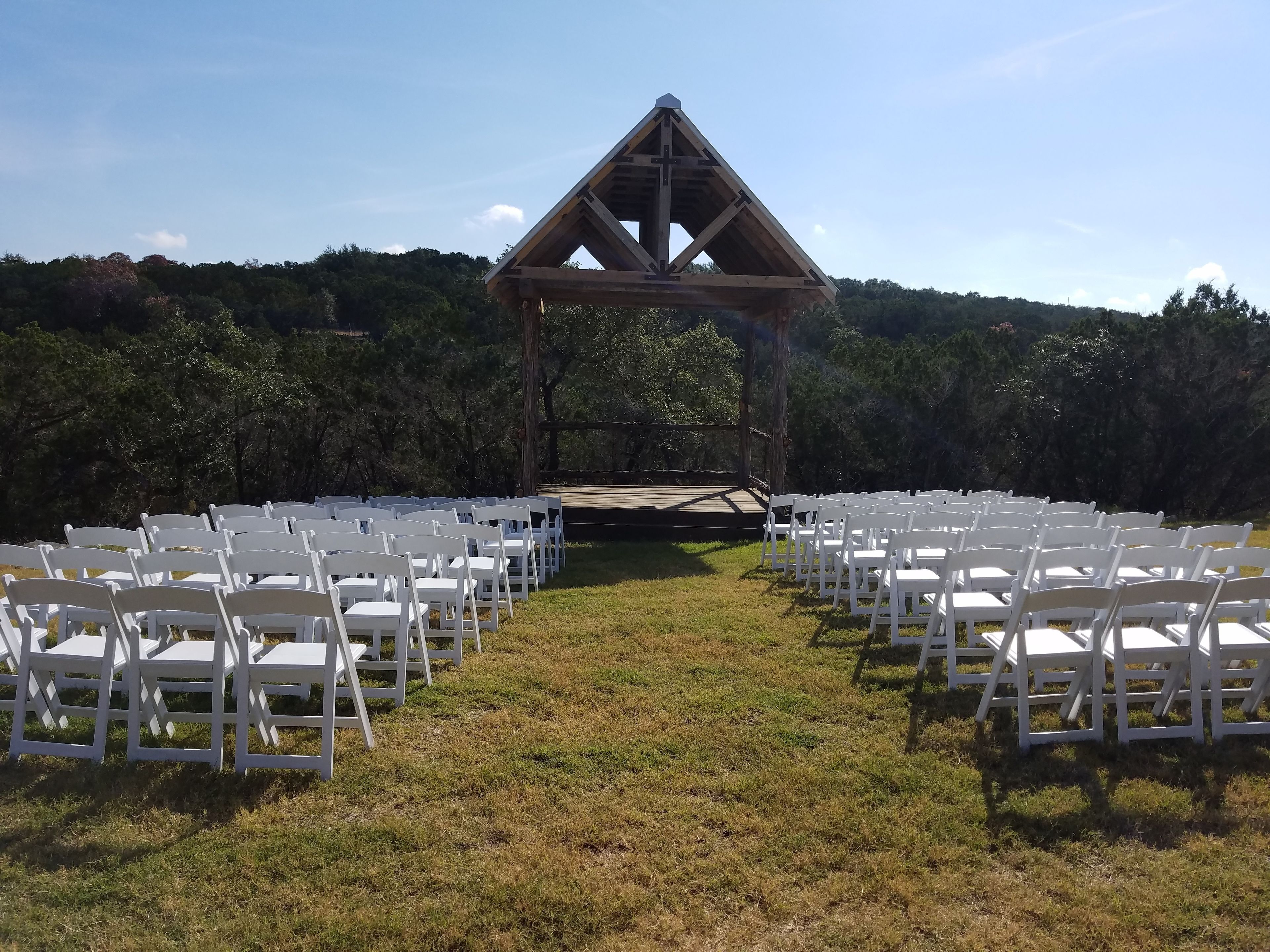 An outdoor wedding venue with white chairs arranged in rows, a wooden stage, and a backdrop of trees.