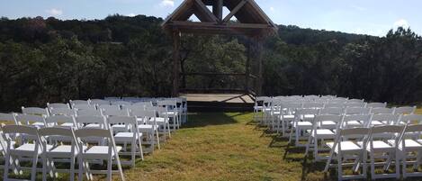 An outdoor wedding venue with white chairs arranged in rows, a wooden stage, and a backdrop of trees.