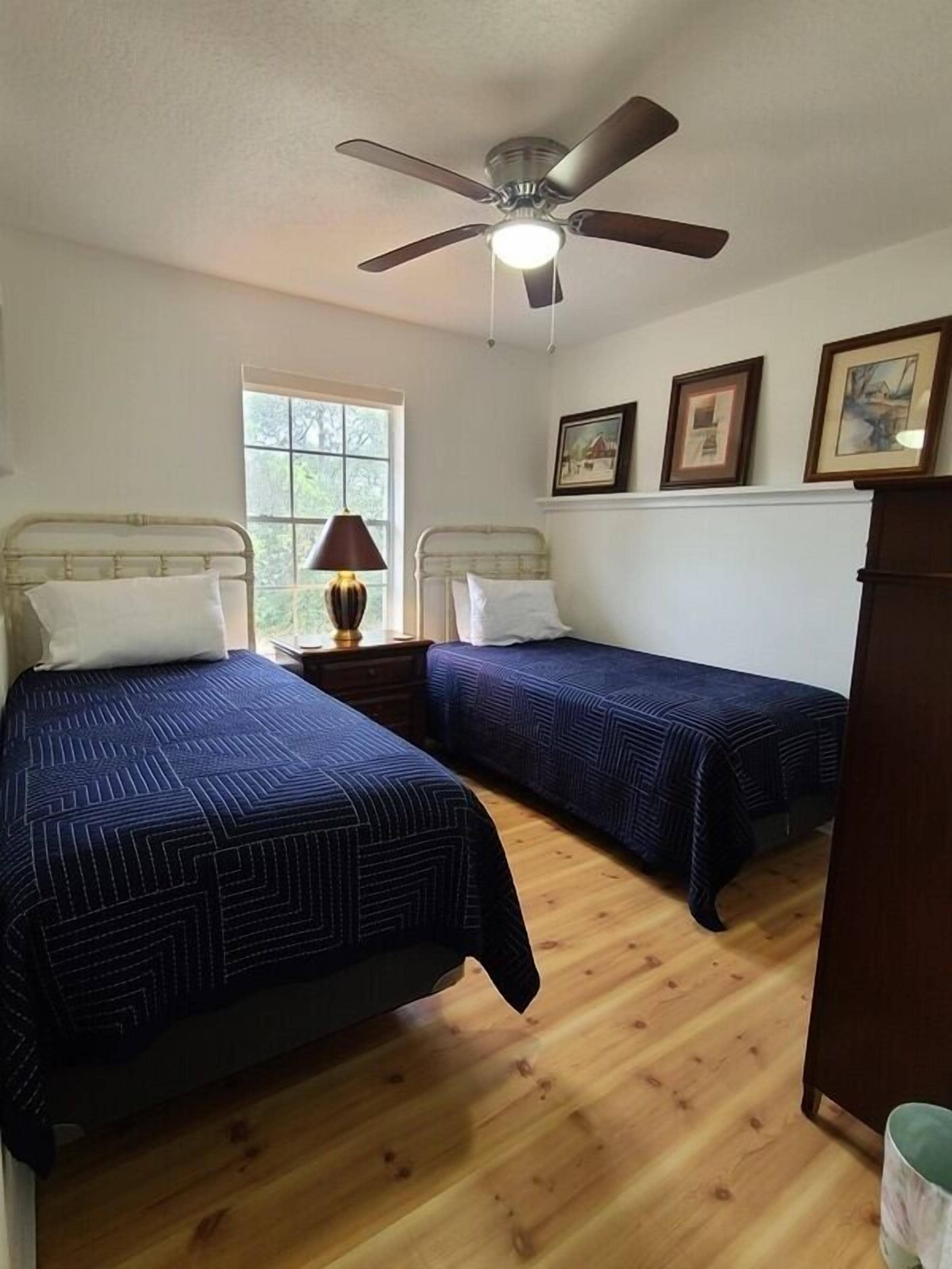 A room with two beds, a ceiling fan, a window, and framed pictures on the wall.