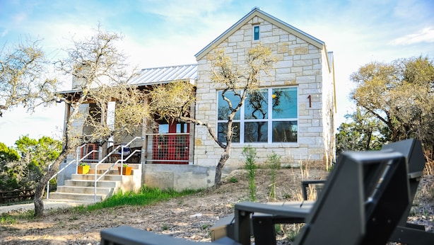 A stone building with a staircase leading to a red door, a tree in front, and a chair in the foreground. - Hill Country Casitas (Dripping Springs)