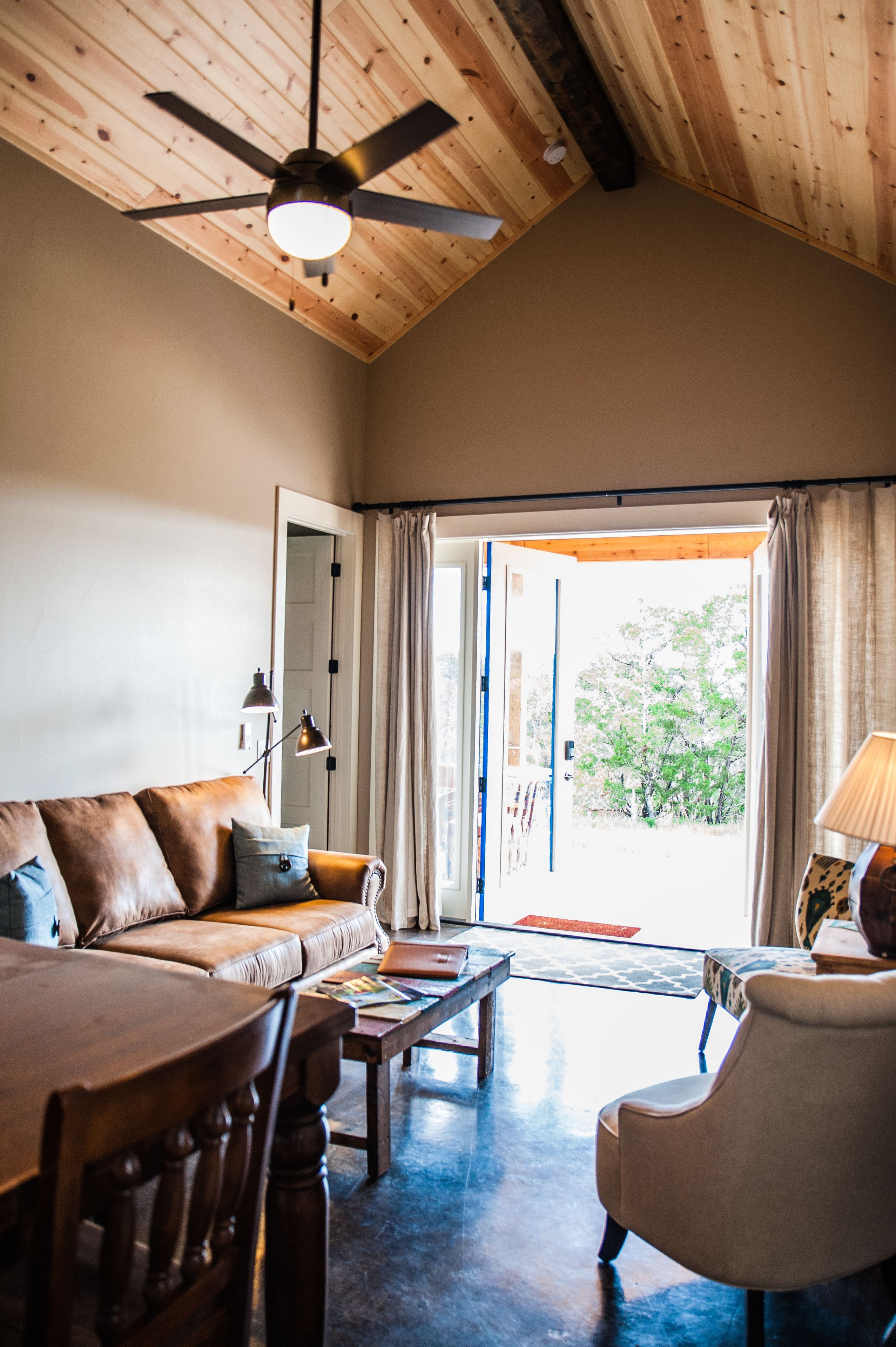 Una sala de estar acogedora con techo de madera, un ventilador de techo, un sofá, una mesa de centro y vistas a un área exterior a través de un ventanal.