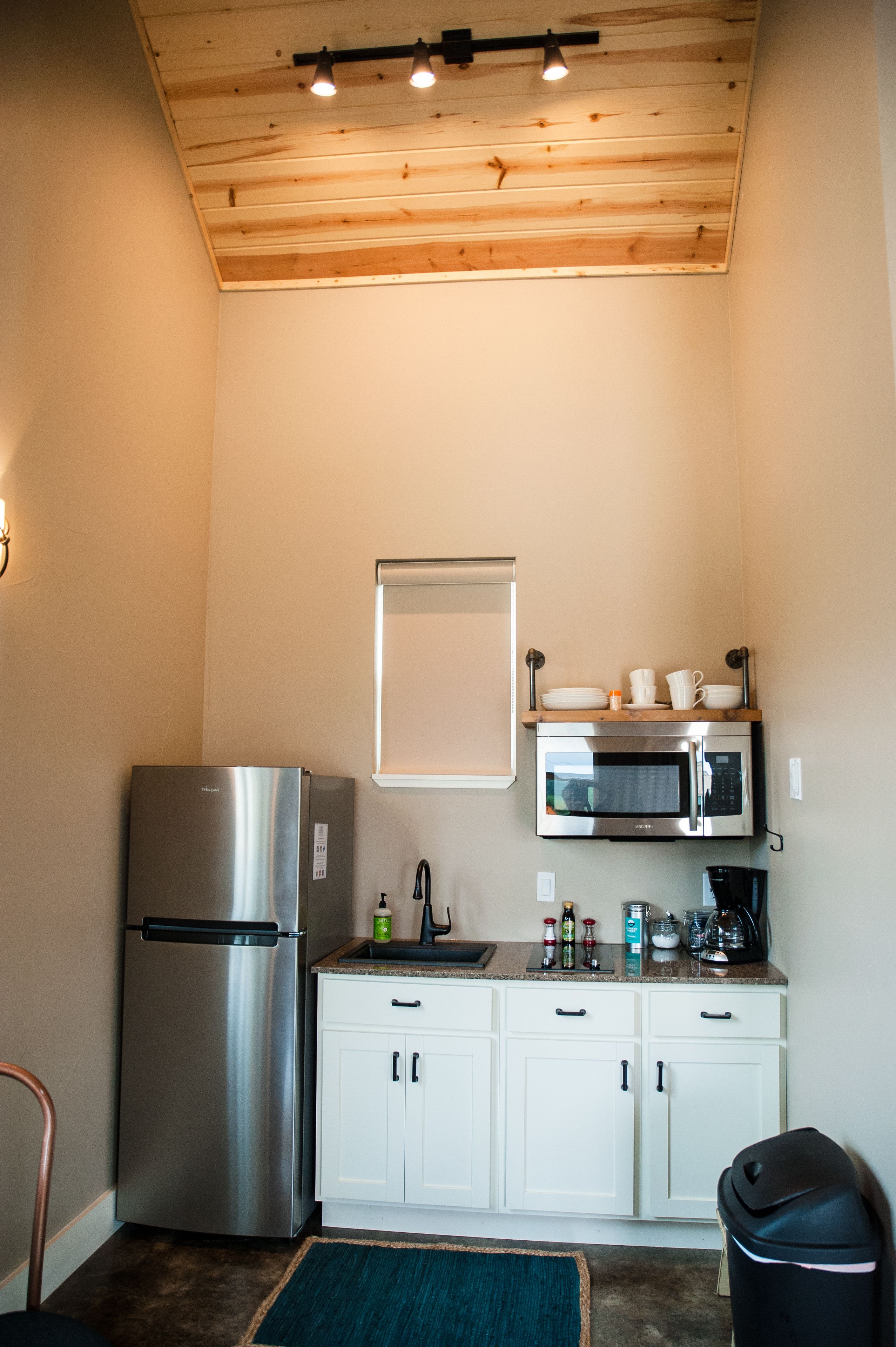 A compact kitchen with a stainless steel refrigerator, a microwave, and a sink. There is a window above the microwave, and a small shelf with dishes.