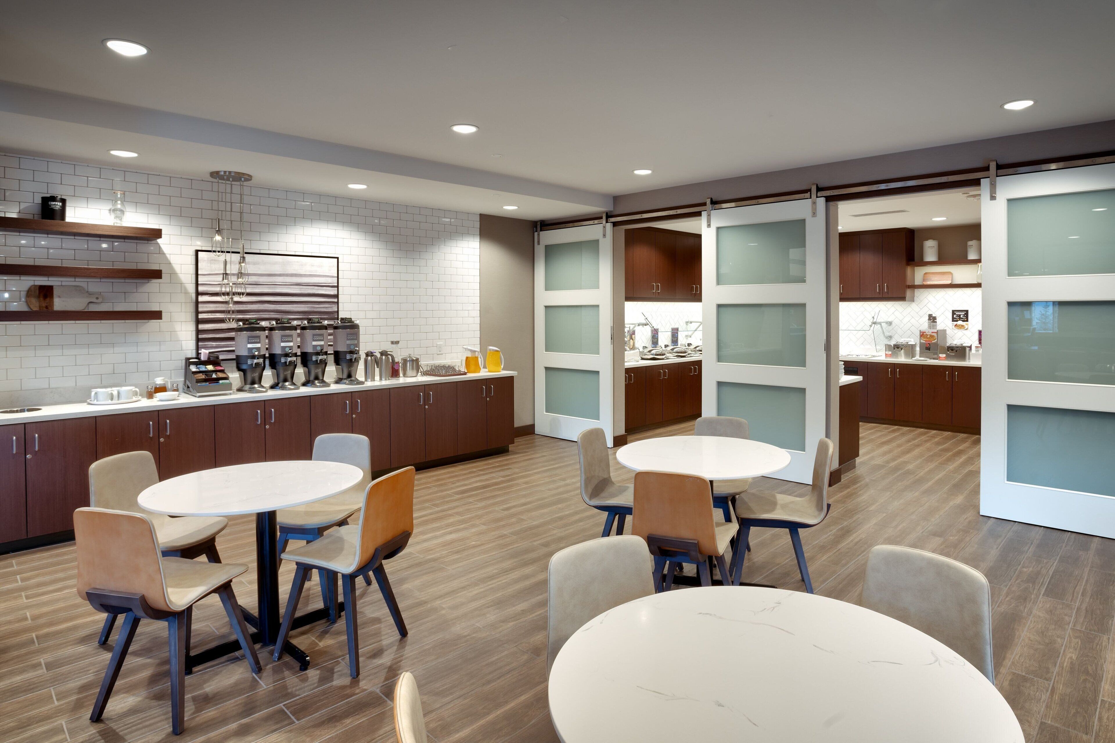 A modern kitchen with white tiled backsplash, wooden cabinets, and a central island with bar tools. There are two round tables with chairs, and a sliding glass door leading to another room.