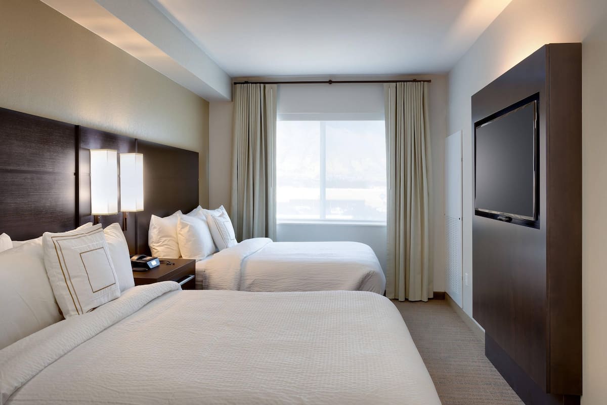 Ein Hotelzimmer mit zwei Betten, einem Fernseher, einem Nachttisch und einem Fenster mit Vorhängen.