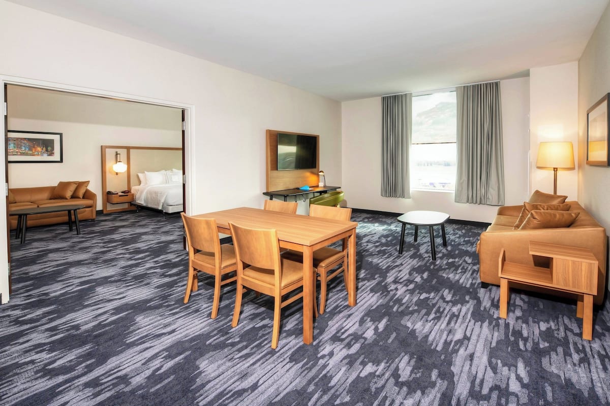 Een hotelkamer met een eettafel, stoelen, een bank, een klein rond tafeltje, een televisie en een raam met gordijnen.
