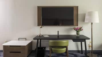 A hotel room with a desk, a chair, a TV, a lamp, a small table with a vase of flowers, and a storage box.