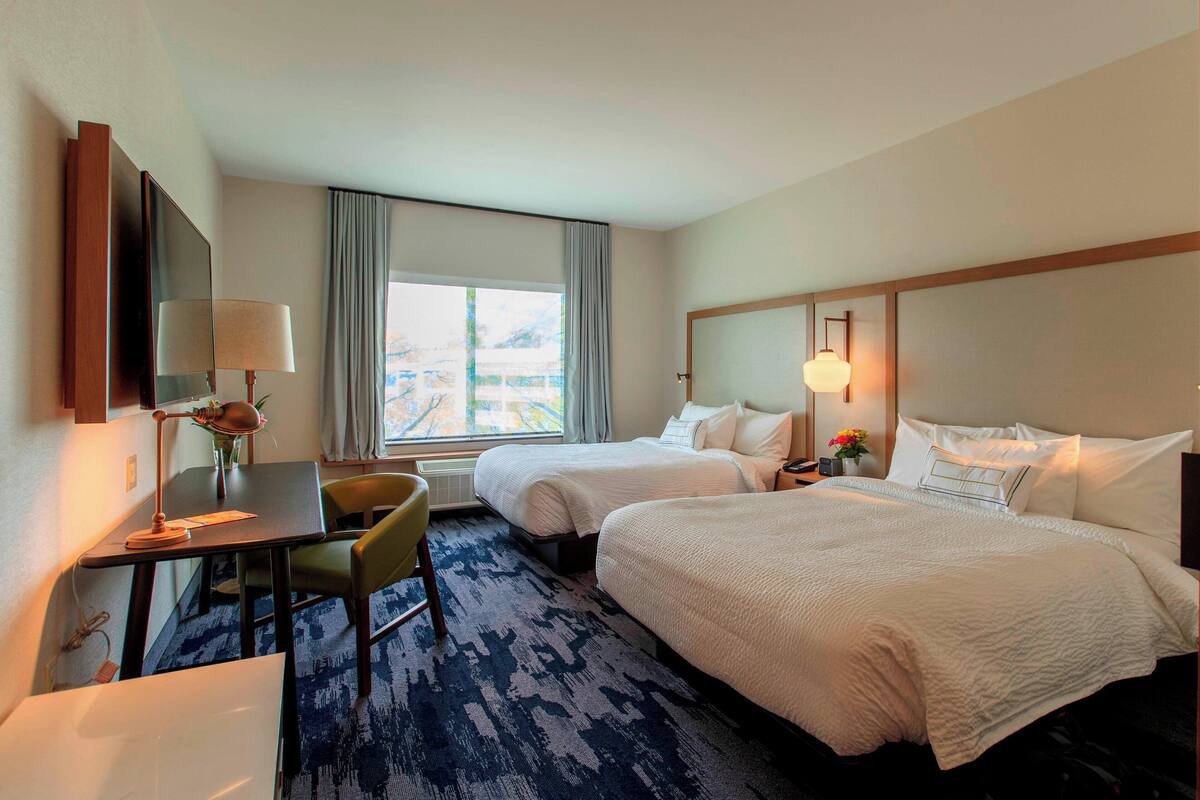 A hotel room with two beds, a desk with a lamp, a chair, a TV, and a large window with curtains.