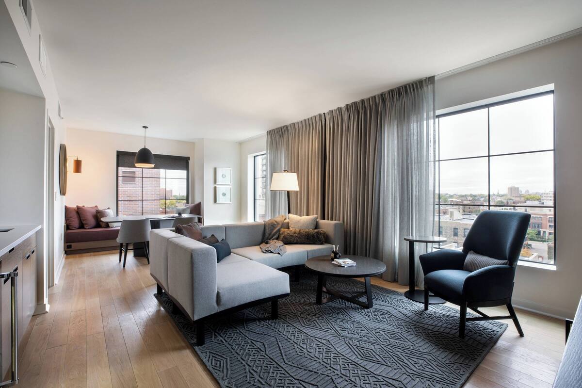 A modern living room with a sofa, armchair, and coffee table, a dining area with a table and chairs, and a large window with curtains.