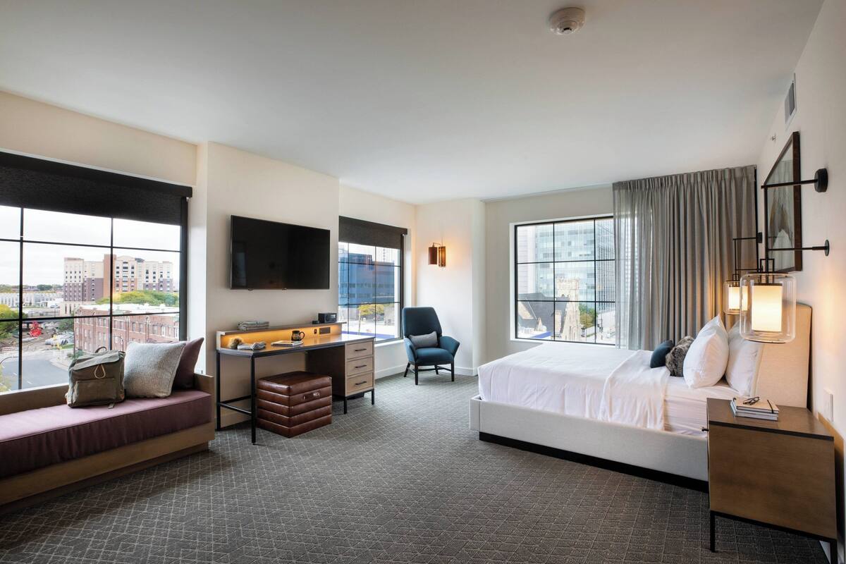 A hotel room with a bed, a desk, a chair, a sofa, and large windows offering a city view.