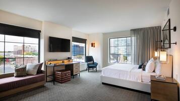 A hotel room with a bed, a desk, a chair, a sofa, and large windows offering a city view.