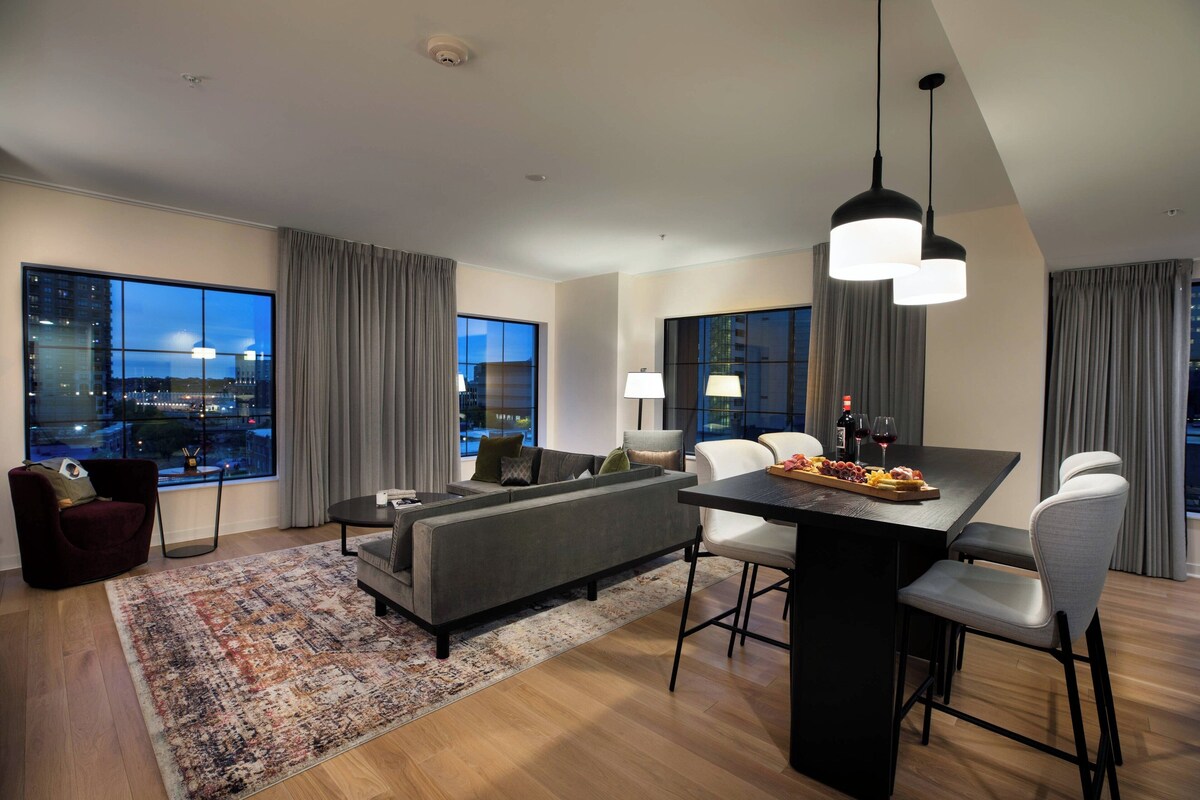 A modern living room with a sofa, dining table, and chairs, large windows with curtains, and a city view.