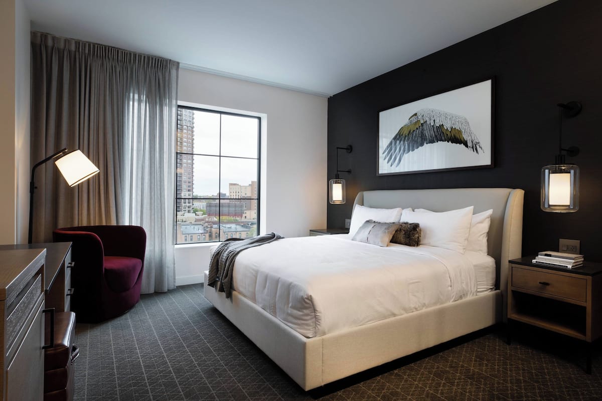 A modern hotel room with a large bed, a red chair, a desk, and a city view through the window.