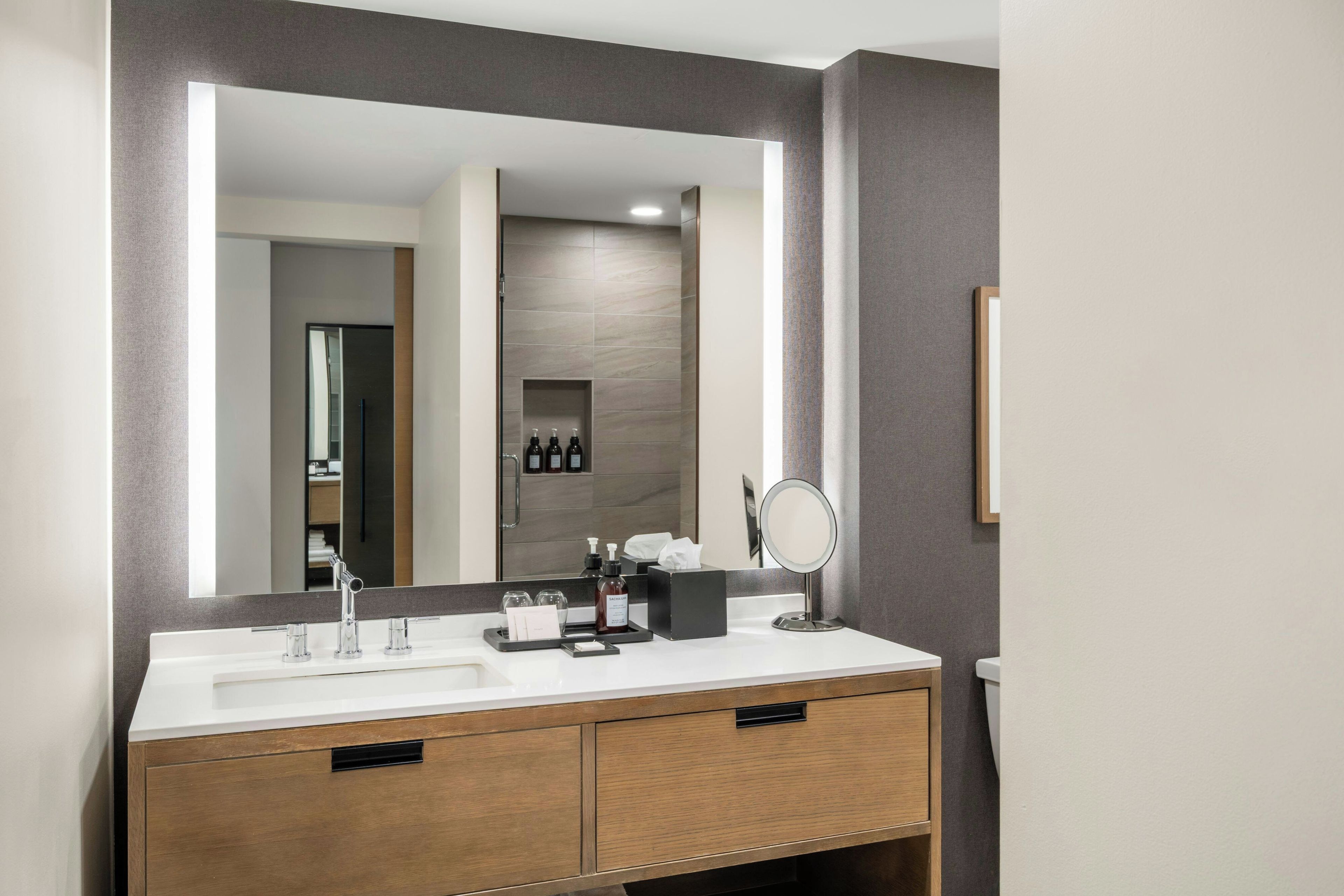 A modern bathroom with a double sink vanity, a large mirror, and a small vanity table with toiletries.