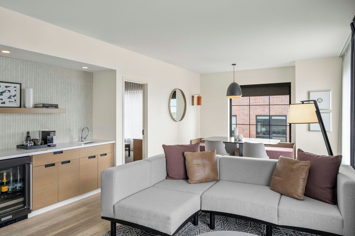 A modern living room with a grey sofa, a kitchen area with a coffee machine, and a view of a brick building through a window.