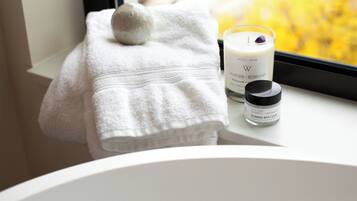 Un baño con una bañera blanca, una toalla blanca, una vela que dice 'LAVANDA + ROMERO' y un frasco de sales de baño.