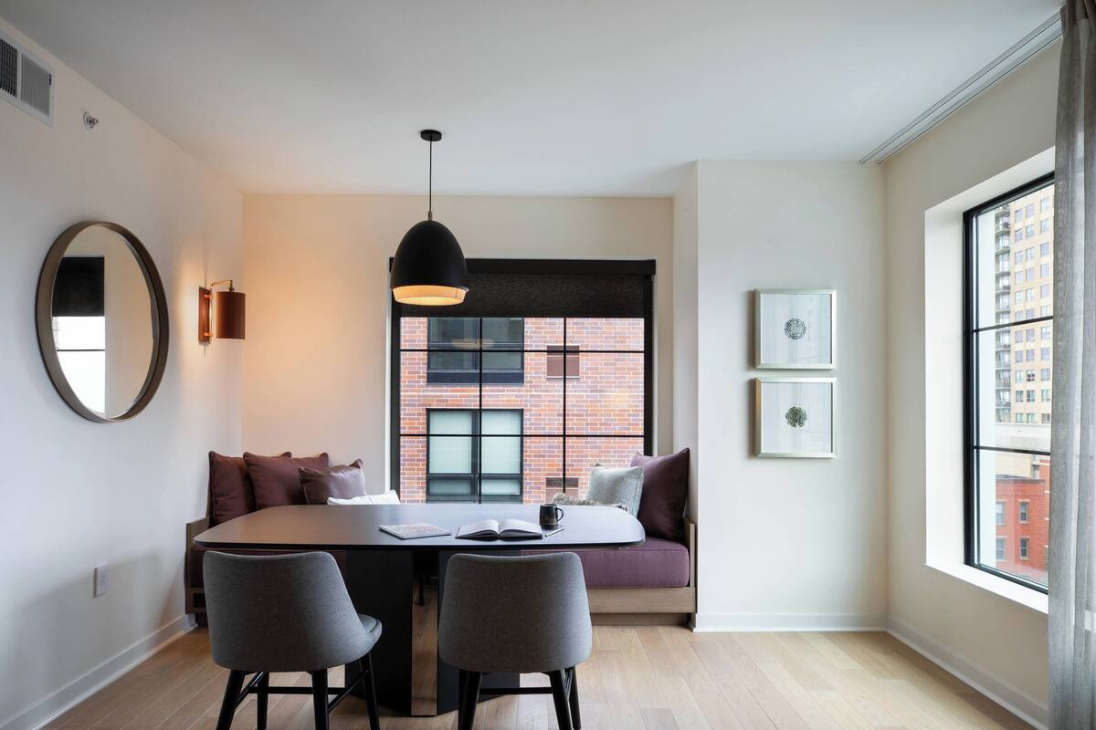 A modern living room with a dining table, two chairs, a sofa, a large window with a view of a brick building, and a pendant light.