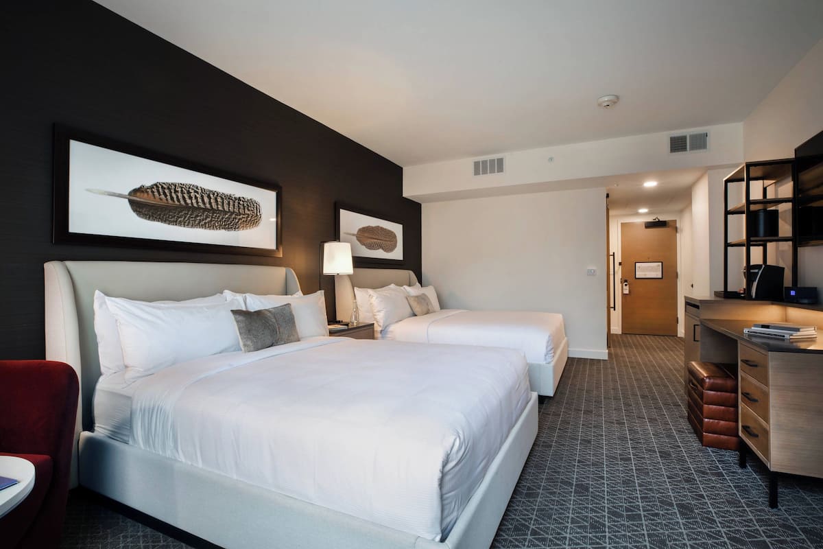 A hotel room with two beds, a desk, and a chair. There are framed pictures on the wall and a lamp on the desk.