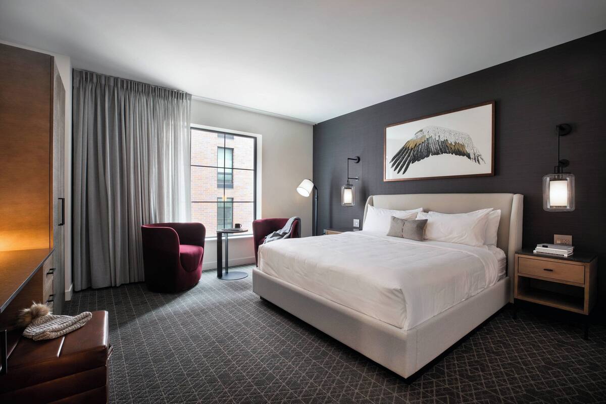 A modern hotel room with a large bed, a desk, a chair, and a window with curtains.